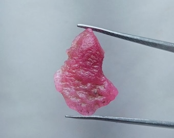 以下が含まれることがあります： ピンセットで保持されたピンクの粗い宝石。宝石は不規則な形をしており、粗くて質感のある表面をしています。