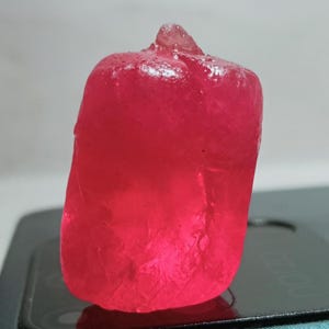 May include: A vibrant, translucent, deep pink gemstone with a rough, irregular, and faceted surface texture. The stone has a rectangular, block-like shape with a small, pointed protrusion at the top, displaying internal light refraction.