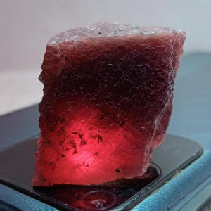 Peut inclure: Une pierre précieuse brute et non taillée, d'un rouge profond virant au rose clair sur le dessus. La pierre présente une surface rugueuse et texturée, semblant être illuminée de l'intérieur, créant un effet de luminescence. Spécimen minéral.
