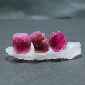 May include: Three raw ruby crystals in varying shapes and sizes, displaying a deep pink to red hue. They are set on a white, irregular-shaped base, with a dark gray background. The crystals have a rough, textured surface.