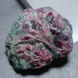 May include: A raw, unpolished mineral specimen featuring a combination of deep red, crystalline ruby sections embedded within a dark, textured green zoisite matrix. The surface displays a mix of rough, matte green areas and shiny red facets.