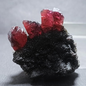 Op de afbeelding: Close-up van een mineraal specimen met helderrode kristallen op een donkere, getextureerde matrix. De kristallen zijn doorschijnend en tonen hun natuurlijke formaties. De compositie benadrukt het contrast tussen de donkere basis en de helderrode kristallen. Natuurlijke formatie.