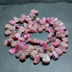 May include: A collection of raw, uncut ruby gemstones in varying shades of pink and red. The gemstones are scattered on a dark surface, showcasing their natural, rough textures and irregular shapes. The arrangement highlights the natural beauty of the stones.