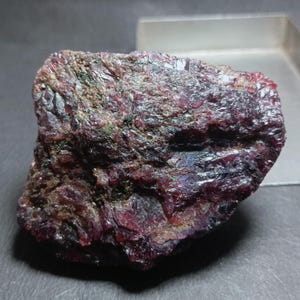 Puede incluir: Un espécimen de rubí en bruto sin pulir que presenta un color rojo oscuro profundo con estructuras cristalinas visibles, texturas superficiales naturales y sutiles inclusiones en la forma irregular y dentada de la piedra mineral.
