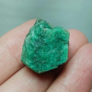 Puede incluir: Una gema de esmeralda verde rugosa con una forma ligeramente irregular. La esmeralda tiene un color verde profundo y rico y una superficie ligeramente texturizada.