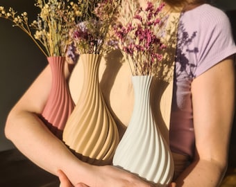 May include: Three ceramic vases in different colors and designs. The vases are filled with dried flowers and are held by a person's hands.