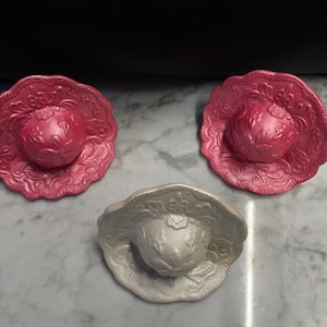 May include: Three decorative hat-shaped objects. Two are a vibrant pink with ornate floral detailing on the brim and crown. The third is a pearlescent white, also with floral patterns. All are on a marble surface.