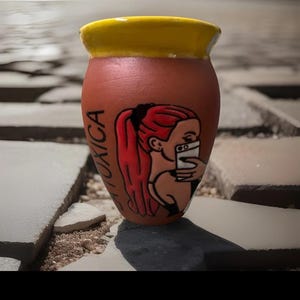 May include: A terracotta-colored ceramic mug with a yellow rim. The mug features a hand-painted illustration of a woman with red hair taking a selfie. The word "TOXICA" is written on the side of the mug.