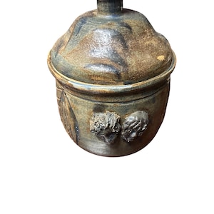 May include: A handcrafted ceramic jar with a lid. The jar is a mix of brown, blue, and tan hues, with a textured surface. Two sculpted faces adorn the side of the jar, adding a unique artistic touch. The jar is a decorative piece.