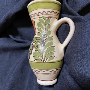 May include: A small, white ceramic pitcher with a green and brown floral design. The pitcher has a handle and a rounded base.