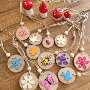 May include: A collection of wooden ornaments featuring floral, insect, and bird designs, strung on twine. The ornaments are made from wooden discs. Several red and white mushroom ornaments are also visible. The text "woodandcraftsbymarie" is present.