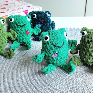 May include: Five crocheted frog plushies, four are green and one is teal. Each frog has large, white eyes and a small ladybug or bee sewn on its chest.