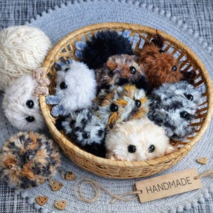 May include: A collection of small, round, fuzzy plush creatures with black button eyes, featuring various fur colors including white, black, brown, and mottled patterns, contained within a round woven wicker basket with a handmade tag.