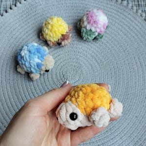 May include: Four small, handmade crochet turtle figurines crafted from soft, plush yarn. Each turtle features a textured, multi-colored shell in shades of yellow, blue, pink, and orange, with a cream-colored body and small black plastic bead eyes.