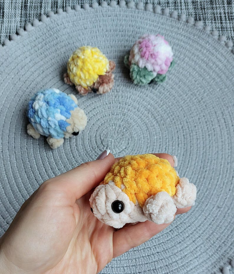 May include: Four small, handmade crochet turtle figurines crafted from soft, plush yarn. Each turtle features a textured, multi-colored shell in shades of yellow, blue, pink, and orange, with a cream-colored body and small black plastic bead eyes.