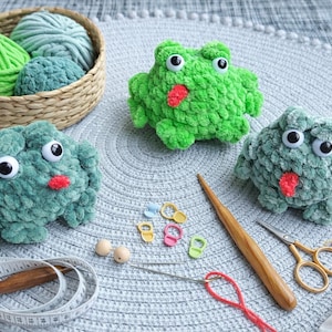 May include: A collection of handmade crochet frog toys in shades of green, with large googly eyes and red accents. The scene includes yarn, crochet hooks, and other crafting supplies, suggesting a DIY project.