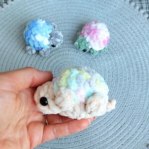 May include: Three small, handmade crochet turtles made from soft, plush yarn. One turtle has a blue and white shell, one has a pink, white, and green shell, and the third features a pastel rainbow shell with a cream-colored body and black eyes.