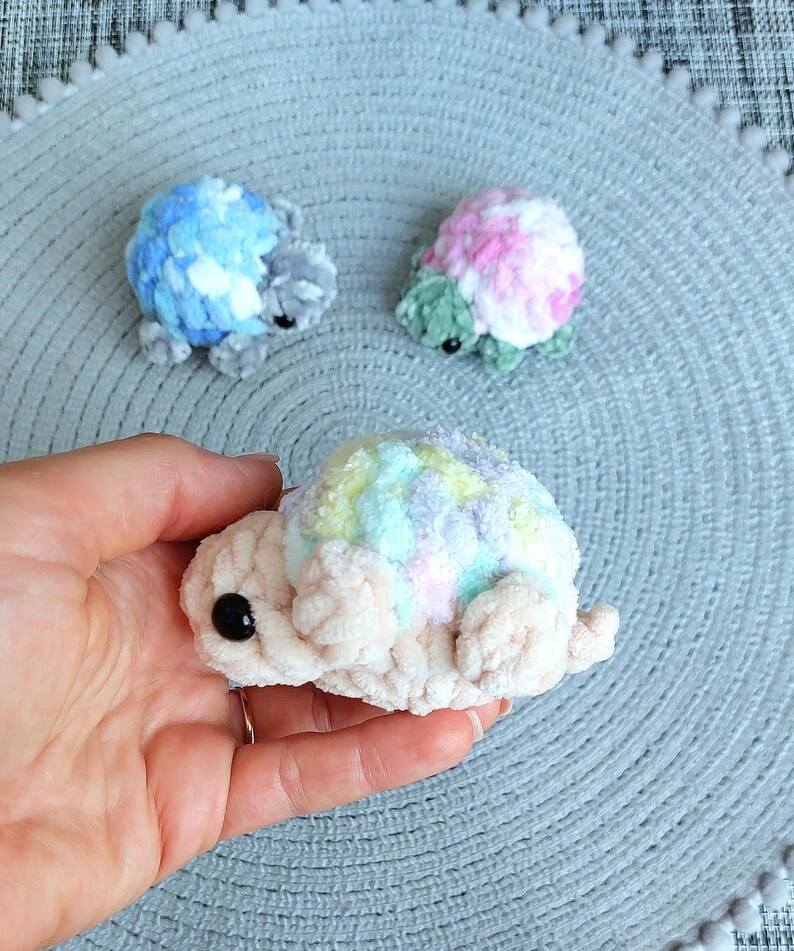 May include: Three small, handmade crochet turtles made from soft, plush yarn. One turtle has a blue and white shell, one has a pink, white, and green shell, and the third features a pastel rainbow shell with a cream-colored body and black eyes.
