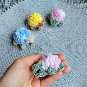 May include: Four small, handmade crochet turtle figurines crafted from soft, plush yarn. Three turtles rest on a textured surface, featuring shells in yellow, pink, and blue, while one pink and green turtle is held in the palm of a hand.