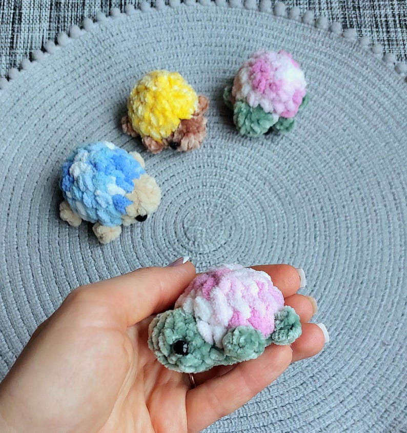 May include: Four small, handmade crochet turtle figurines crafted from soft, plush yarn. Three turtles rest on a textured surface, featuring shells in yellow, pink, and blue, while one pink and green turtle is held in the palm of a hand.