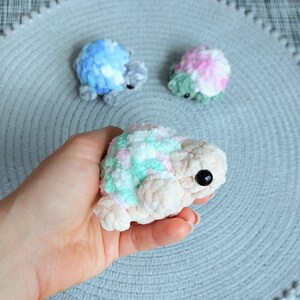 May include: Three small, plush, crocheted turtle figures with rounded shells. One turtle is held, featuring a cream-colored body and a shell with pastel pink, green, and white yarn. Two additional turtles in blue and pink-green sit nearby.