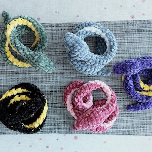 Könnte beinhalten: Fünf gehäkelte Schlangen-Spielzeuge in verschiedenen Farben: Grün mit gelben Streifen, Blau mit Weiß, Lila mit Gelb, Schwarz mit Gelb und Rosa mit Weiß. Die Schlangen sind auf einer grauen, strukturierten Oberfläche angeordnet.