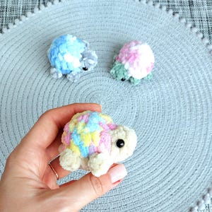 May include: Three small, handmade crochet turtles crafted from soft, plush yarn. One turtle features a variegated shell of pastel pink, yellow, and blue with a cream-colored body. Two additional turtles in blue and pink patterns sit in the background.