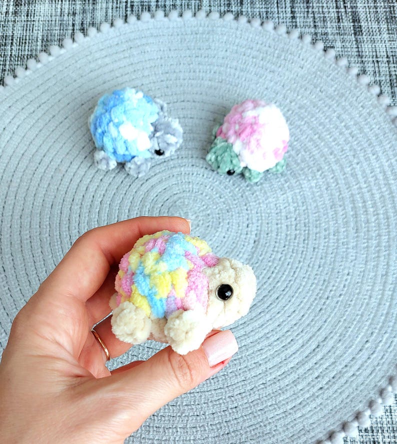 May include: Three small, handmade crochet turtles crafted from soft, plush yarn. One turtle features a variegated shell of pastel pink, yellow, and blue with a cream-colored body. Two additional turtles in blue and pink patterns sit in the background.