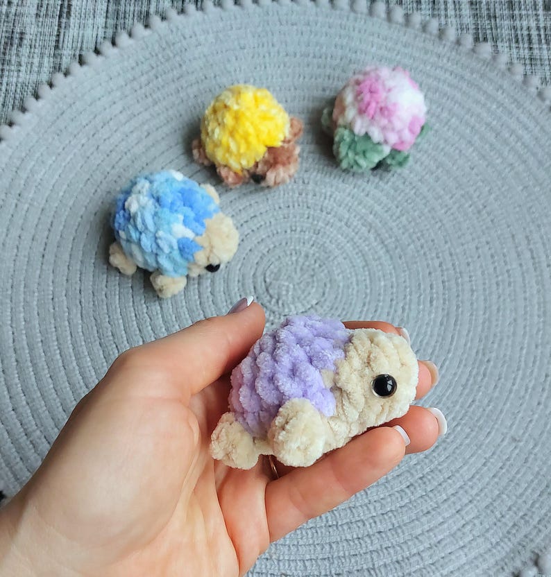 May include: Four small, plush, crocheted turtle figures with soft, textured yarn shells in various colors including yellow, blue, pink, and purple. Each turtle has a cream-colored head and limbs with a single black bead eye on each side.