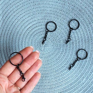 May include: Four black metal keychains, each consisting of a circular split ring attached to a short chain and a small lobster claw clasp. The keychains have a matte black finish and are arranged on a textured light blue surface.