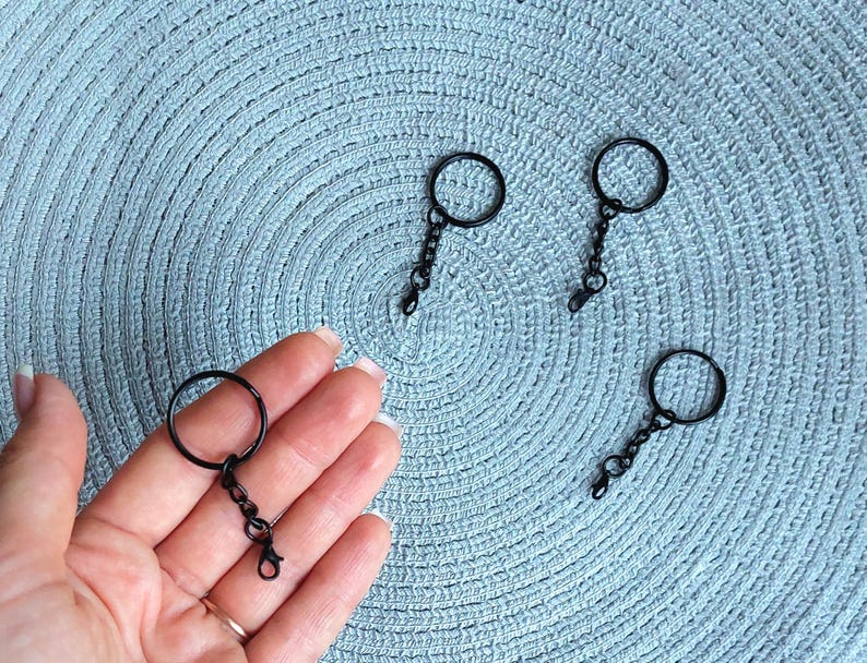 May include: Four black metal keychains, each consisting of a circular split ring attached to a short chain and a small lobster claw clasp. The keychains have a matte black finish and are arranged on a textured light blue surface.