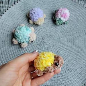 May include: Four small, plush, crocheted turtle figures made from soft yarn. One turtle is yellow and brown, one is light purple and beige, one is mint green and beige, and one is a variegated mix of pink, white, and green with beige accents.