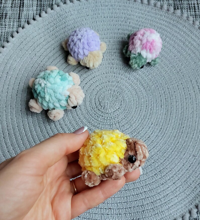May include: Four small, plush, crocheted turtle figures made from soft yarn. One turtle is yellow and brown, one is light purple and beige, one is mint green and beige, and one is a variegated mix of pink, white, and green with beige accents.