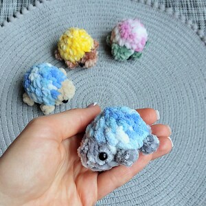 May include: Four small, handmade crochet turtle figurines made from soft, plush yarn. One is held in the foreground, featuring blue, white, and gray tones. Three others in the background are yellow and brown, blue and white, and pink and green.