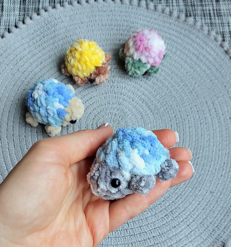 May include: Four small, handmade crochet turtle figurines made from soft, plush yarn. One is held in the foreground, featuring blue, white, and gray tones. Three others in the background are yellow and brown, blue and white, and pink and green.