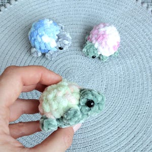 May include: Three small, plush, crocheted turtle figurines made from soft, fuzzy yarn. One turtle has a blue and white shell, one has a pink and white shell, and the one held in the hand features a pale green and soft pink shell with dark green limbs.