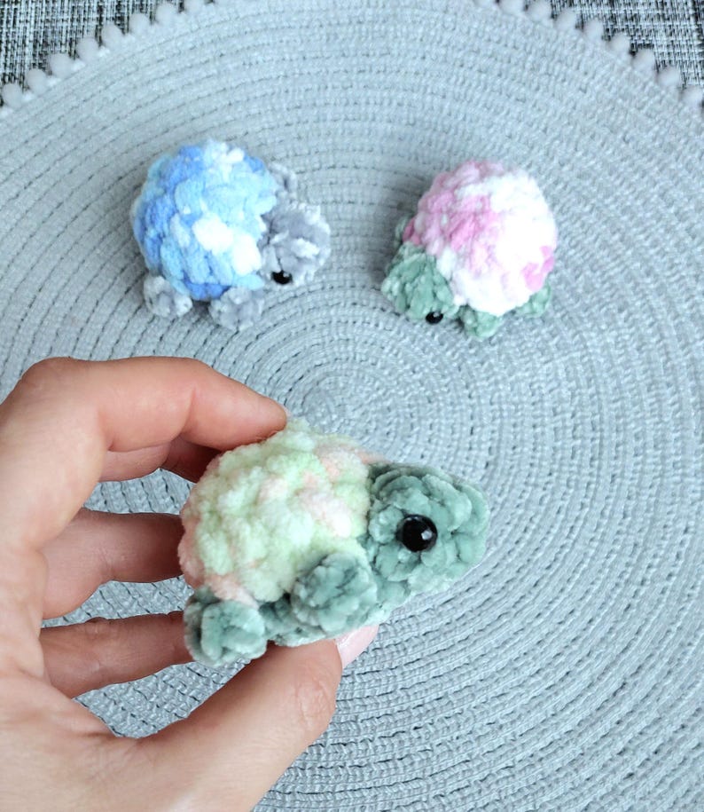 May include: Three small, plush, crocheted turtle figurines made from soft, fuzzy yarn. One turtle has a blue and white shell, one has a pink and white shell, and the one held in the hand features a pale green and soft pink shell with dark green limbs.