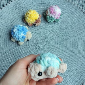 May include: Four small, plush, crocheted turtle figures with soft, textured yarn shells in various colors including yellow, pink, blue, and teal. Each turtle features a light beige head and limbs with small, round, black plastic eyes.