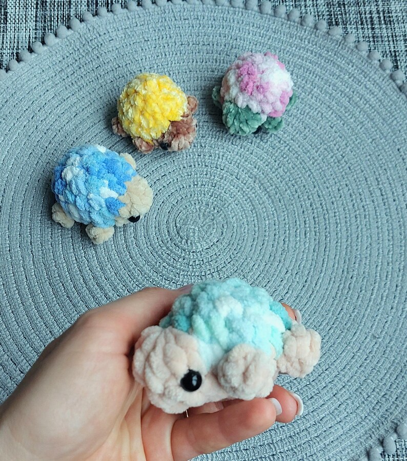 May include: Four small, plush, crocheted turtle figures with soft, textured yarn shells in various colors including yellow, pink, blue, and teal. Each turtle features a light beige head and limbs with small, round, black plastic eyes.