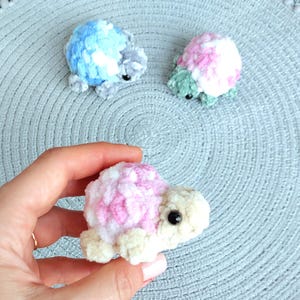 May include: Three small, handmade crochet turtle figurines crafted from soft, plush yarn. One turtle features a blue and white shell, another has a pink and white shell with green limbs, and the third has a pink and white shell with cream limbs.