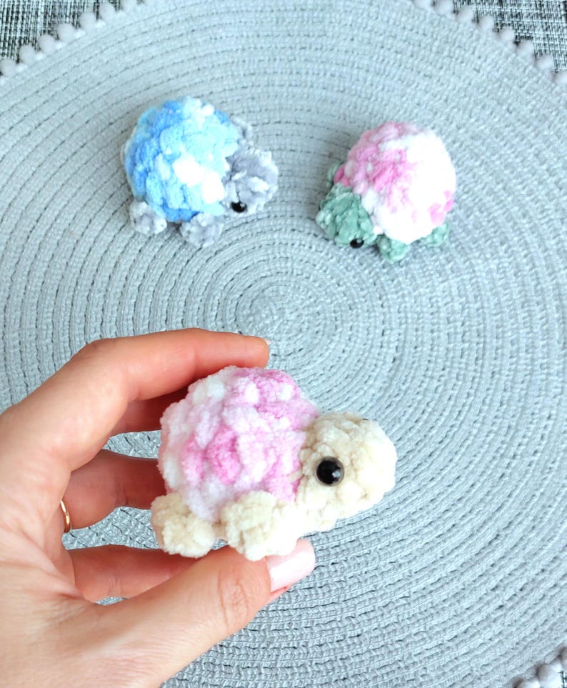 May include: Three small, handmade crochet turtle figurines crafted from soft, plush yarn. One turtle features a blue and white shell, another has a pink and white shell with green limbs, and the third has a pink and white shell with cream limbs.