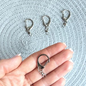 May include: Four silver-toned metal lobster claw clasps with swivel connectors. Each clasp features a rounded D-shaped loop and a spring-loaded gate mechanism for secure attachment, designed for use in jewelry making or as keychain hardware components.