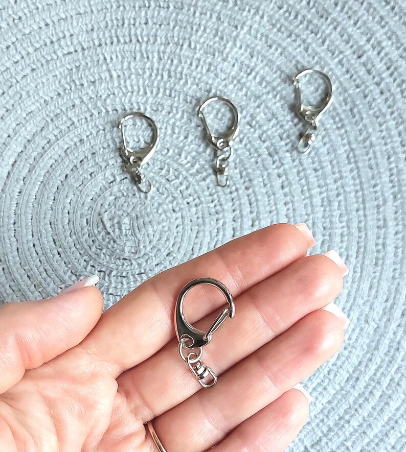 May include: Four silver-toned metal lobster claw clasps with swivel connectors. Each clasp features a rounded D-shaped loop and a spring-loaded gate mechanism for secure attachment, designed for use in jewelry making or as keychain hardware components.