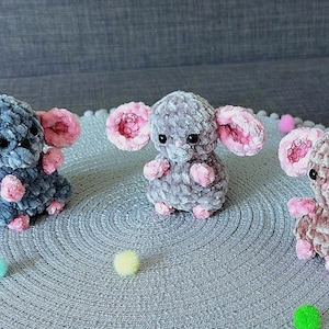 May include: Three handmade crochet mice in shades of gray and brown, each with large pink-lined ears and small pink paws. They are sitting on a gray circular mat with colorful pom-poms scattered around.
