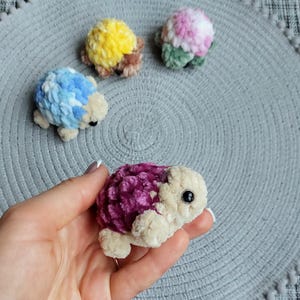 May include: Four small, plush, crocheted turtle figurines with soft, textured shells in vibrant colors including yellow, blue, pink, and deep purple. Each turtle features a cream-colored head, small limbs, and a single black bead eye on each side.