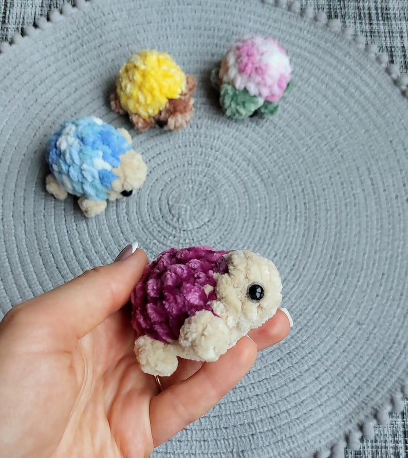 May include: Four small, plush, crocheted turtle figurines with soft, textured shells in vibrant colors including yellow, blue, pink, and deep purple. Each turtle features a cream-colored head, small limbs, and a single black bead eye on each side.