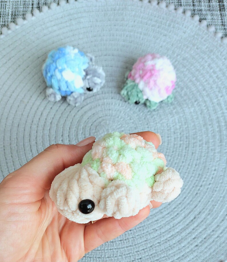 May include: Three small, handmade crochet turtles crafted from soft, plush yarn. One turtle features a cream-colored body with a pastel green and peach shell, while the others display blue and white or pink and green color combinations.