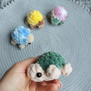 May include: Four small, handmade crochet turtle figurines made from soft, plush yarn. One turtle is held in the foreground with a dark green shell and cream-colored body. Three others in blue, yellow, and pink-green sit in the background.