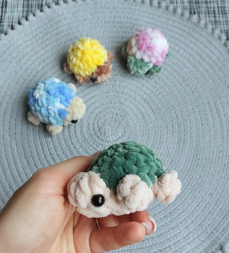 May include: Four small, handmade crochet turtle figurines made from soft, plush yarn. One turtle is held in the foreground with a dark green shell and cream-colored body. Three others in blue, yellow, and pink-green sit in the background.