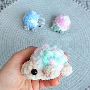 May include: Three small, plush, crocheted turtle figures. One is held in the foreground, featuring a beige head and limbs with a pastel pink, white, and mint green shell. Two smaller turtles in blue and pink sit in the background.